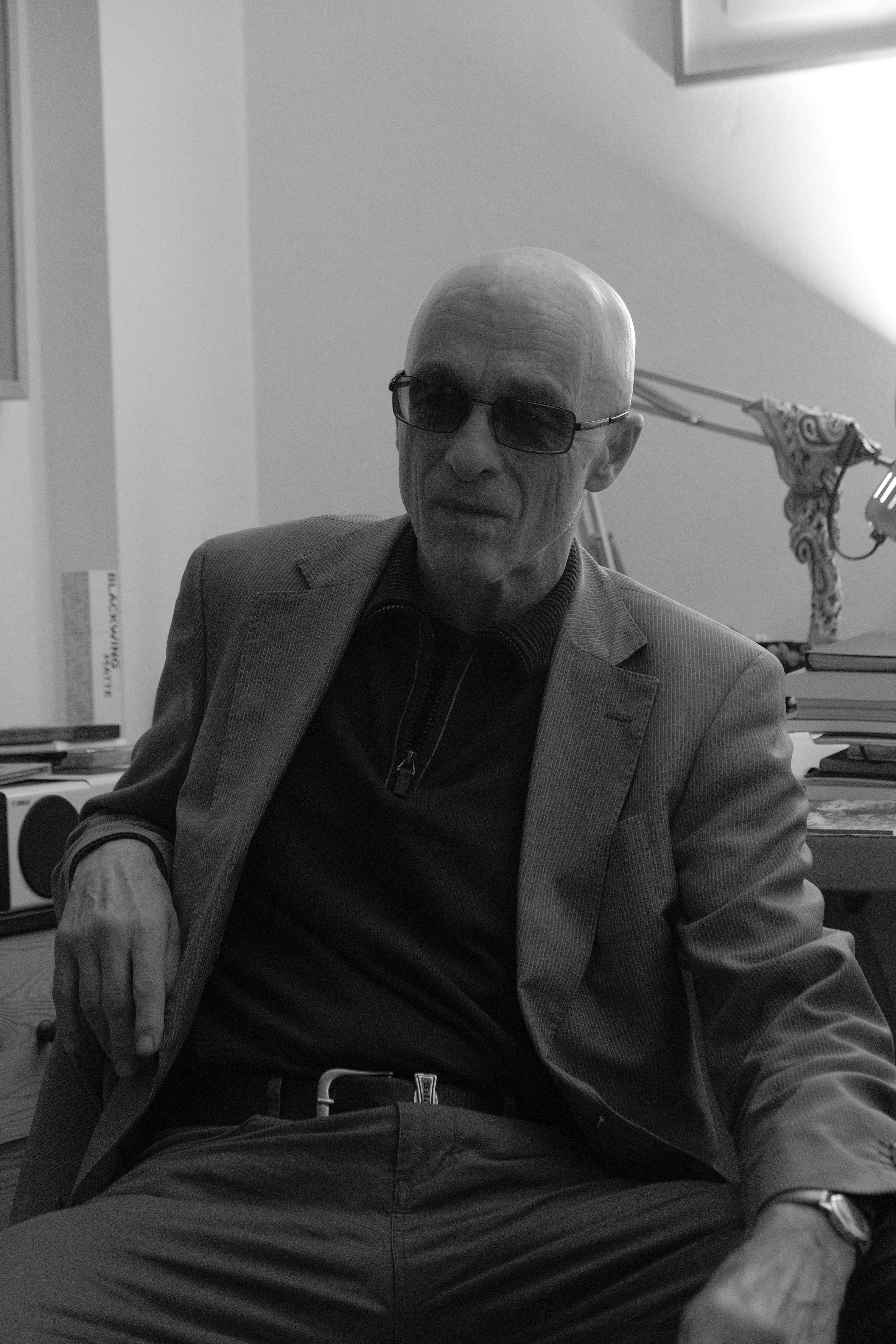 Black and white portrait of a senior man in sunglasses, seated indoors in a modern setting.