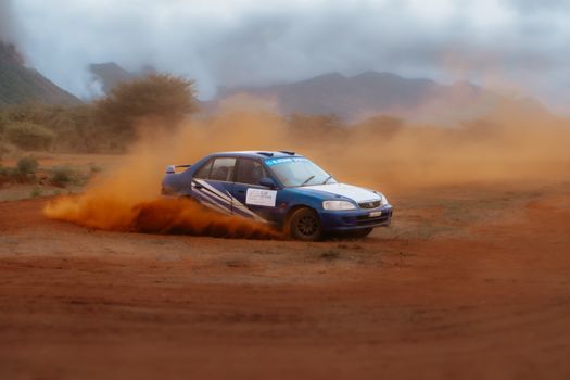 Blue rally car executing a sharp turn in a dusty, rugged outdoor setting.