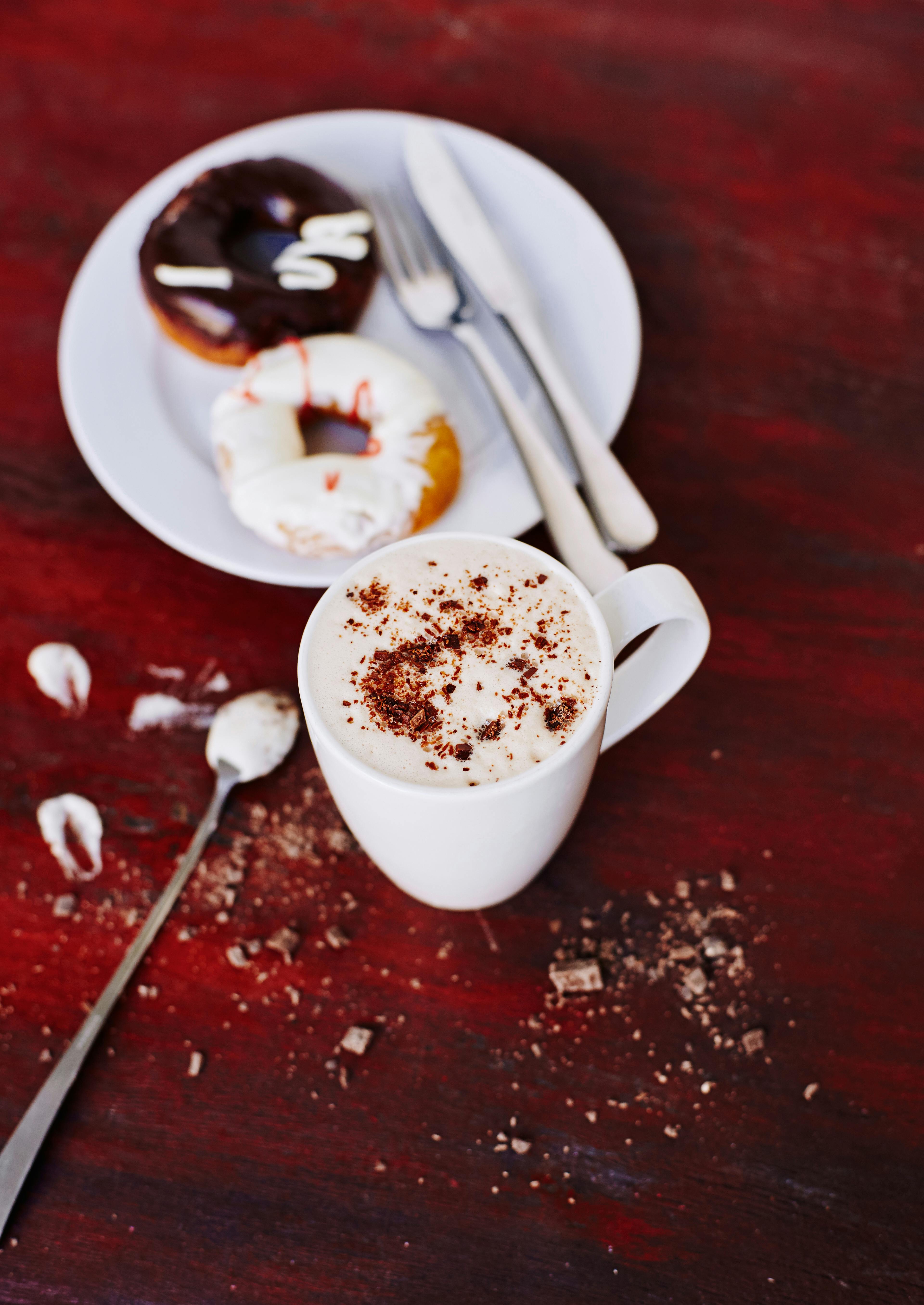 Cup of coffee with foam and two donuts on a white plate on a red rustic surface.