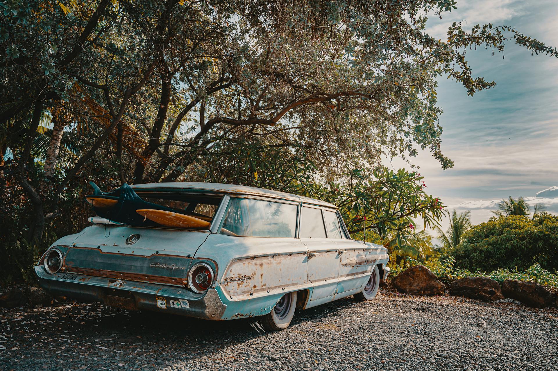 A classic rusty car with surfboards parked outdoors on a gravel road surrounded by fall trees.