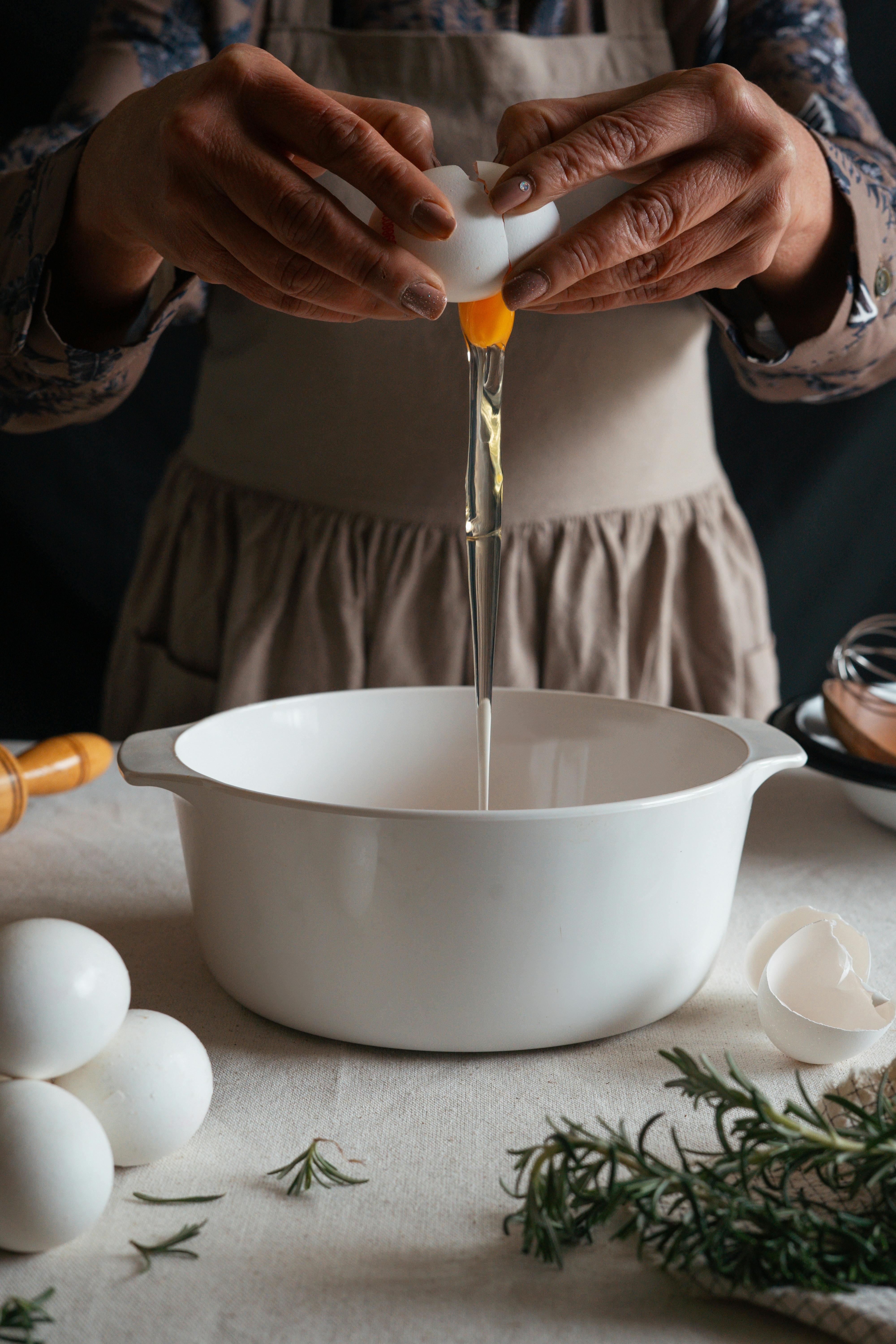 Hands breaking an egg into a white mixing bowl with fresh rosemary on table.
