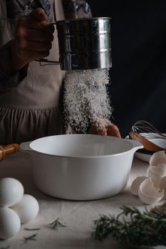 A person sifting flour into a white bowl with eggshells and herbs nearby, creating a rustic kitchen scene.