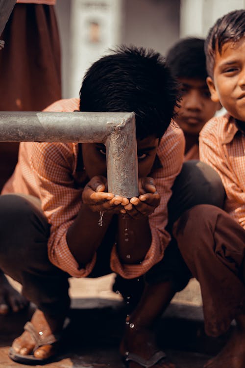 Free Ethnic boys in similar clothes sitting on haunches and drinking water from pipe on town street Stock Photo