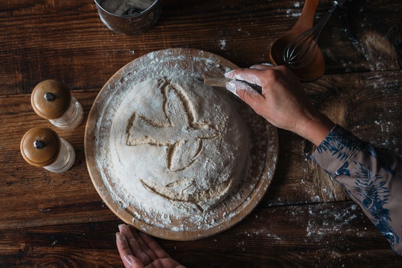 A person prepares rustic homemade bread dough with flour on a wooden table, creating a cozy atmosphere.