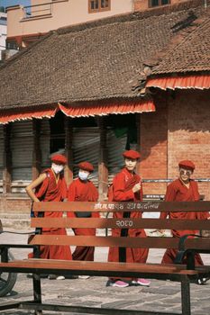 Four Buddhist monks in red robes walk past traditional Nepalese architecture.
