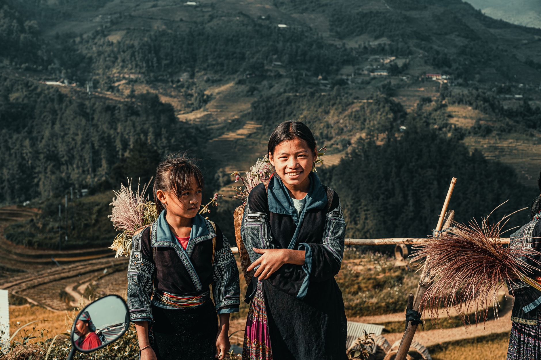 Two ethnic girls in traditional attire with mountainous background, smiling.