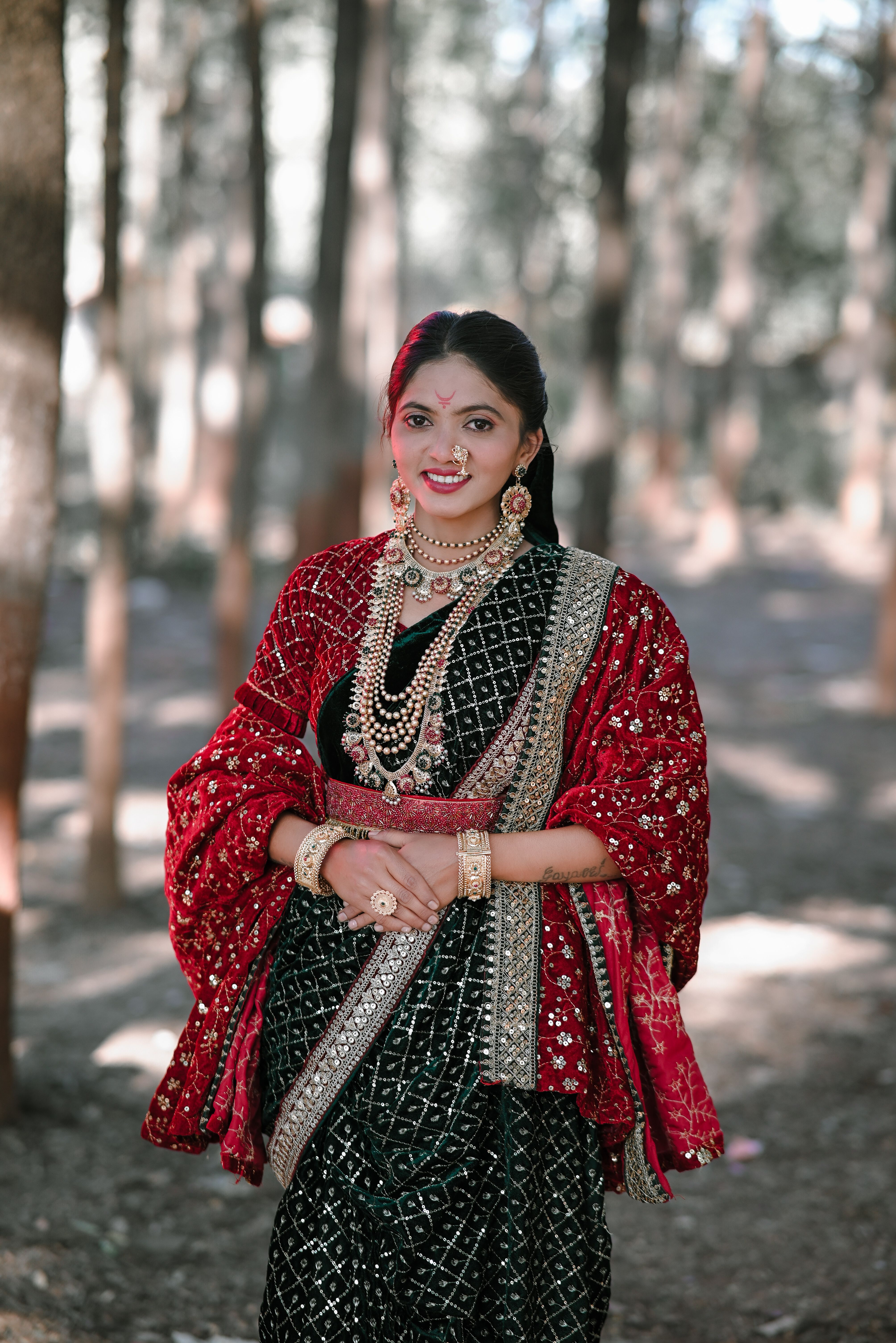 A woman in vibrant traditional Indian clothing stands gracefully outdoors with a forest background.