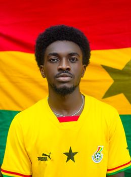 Man wearing Ghana jersey in front of Ghana flag. Vibrant colors.