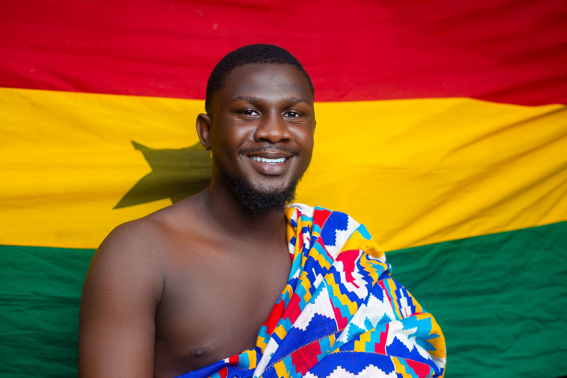 Smiling man in vibrant Kente cloth against Ghana flag background.