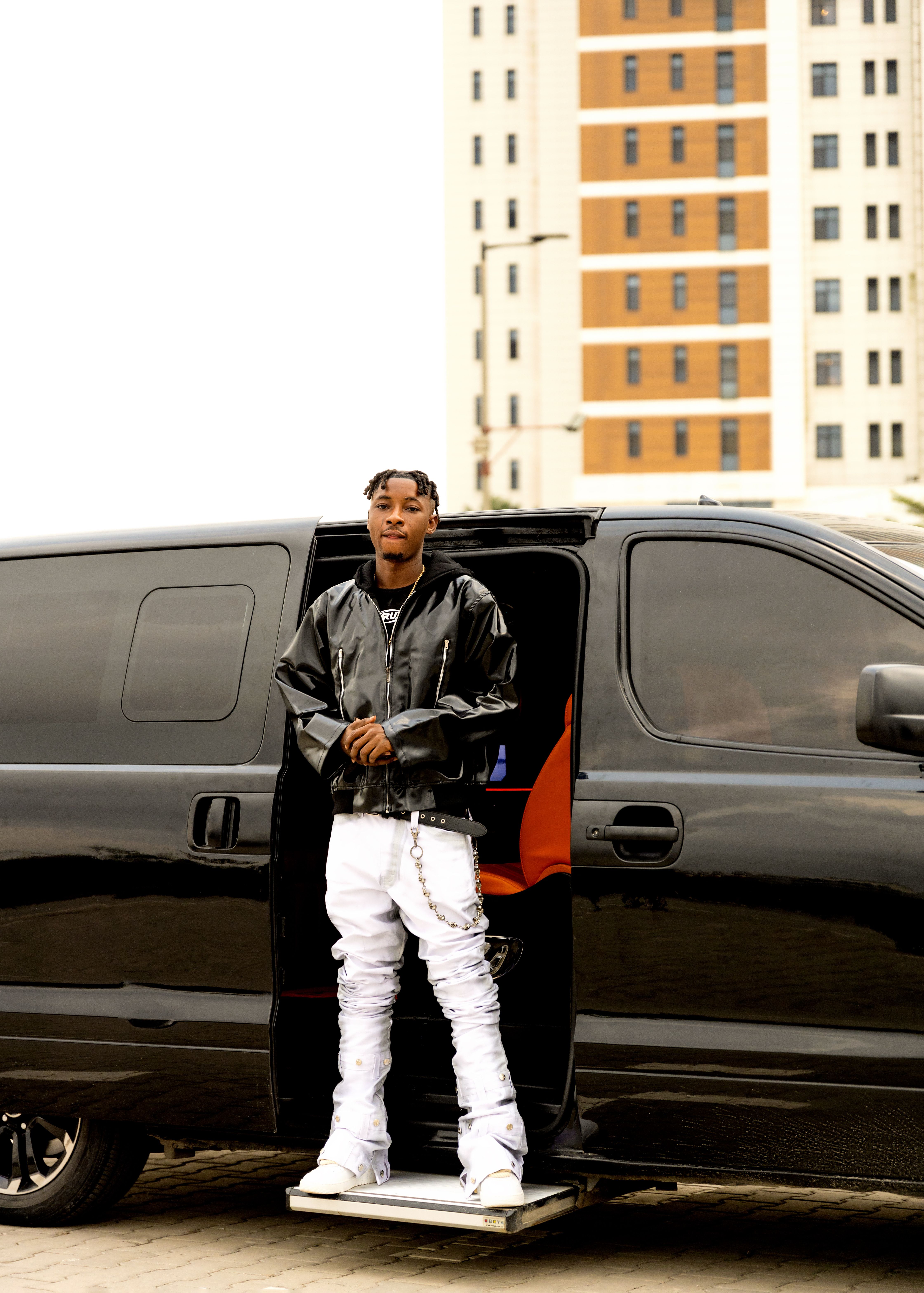 A fashionable young man in black jacket and white pants standing by a black van with urban background.