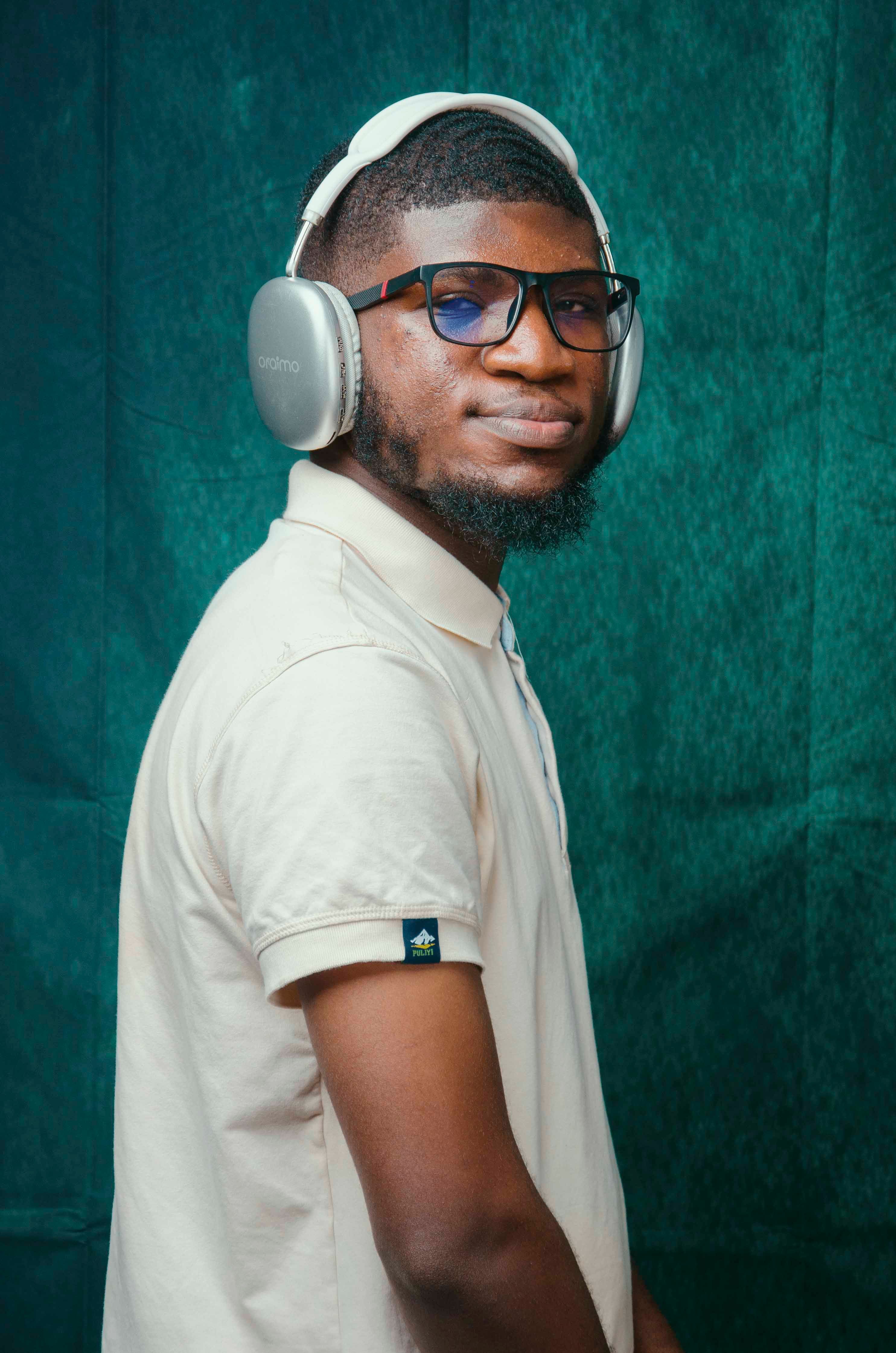 A man wearing headphones and glasses poses confidently against a teal backdrop in a studio.