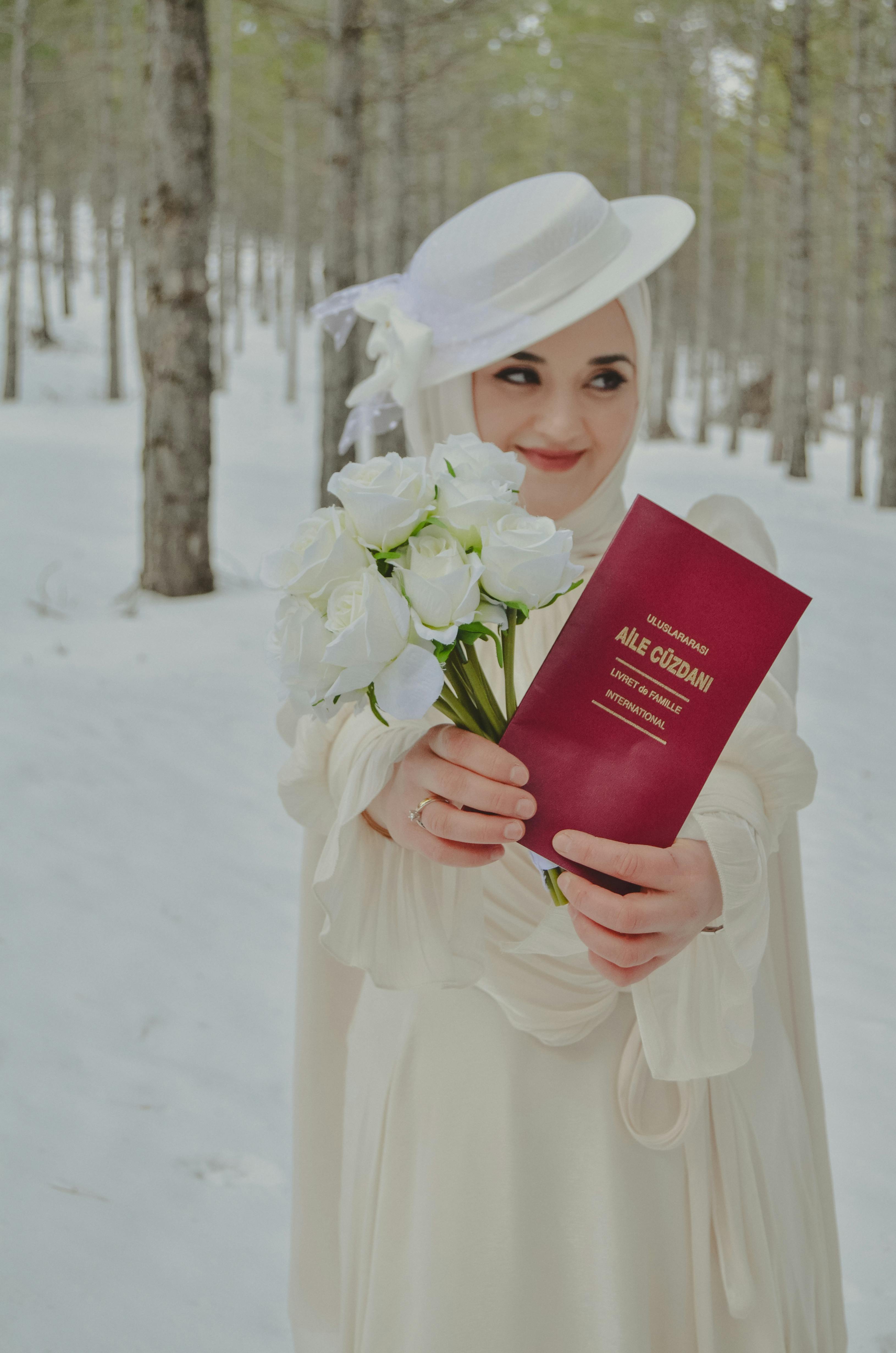 A bride in snow, wearing a white dress and hat, holding a certificate and bouquet.