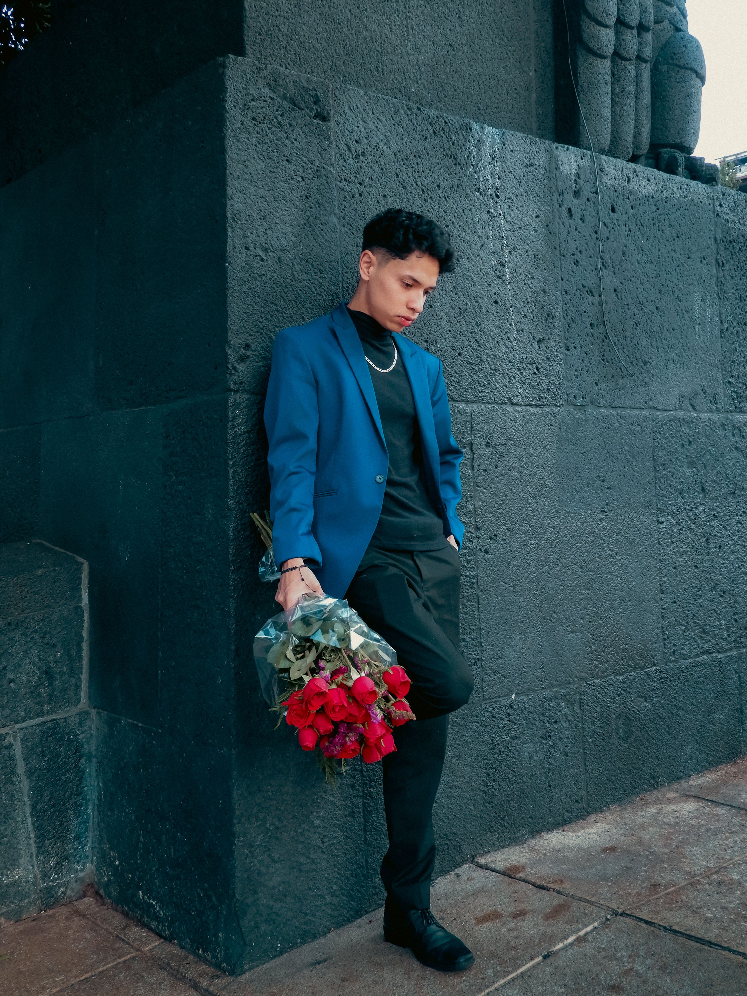 Stylish man in blue blazer with roses, leaning against urban wall, conveying emotion.