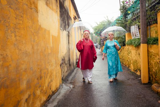 Two women in traditional attire walking with umbrellas in Hội An, Vietnam.