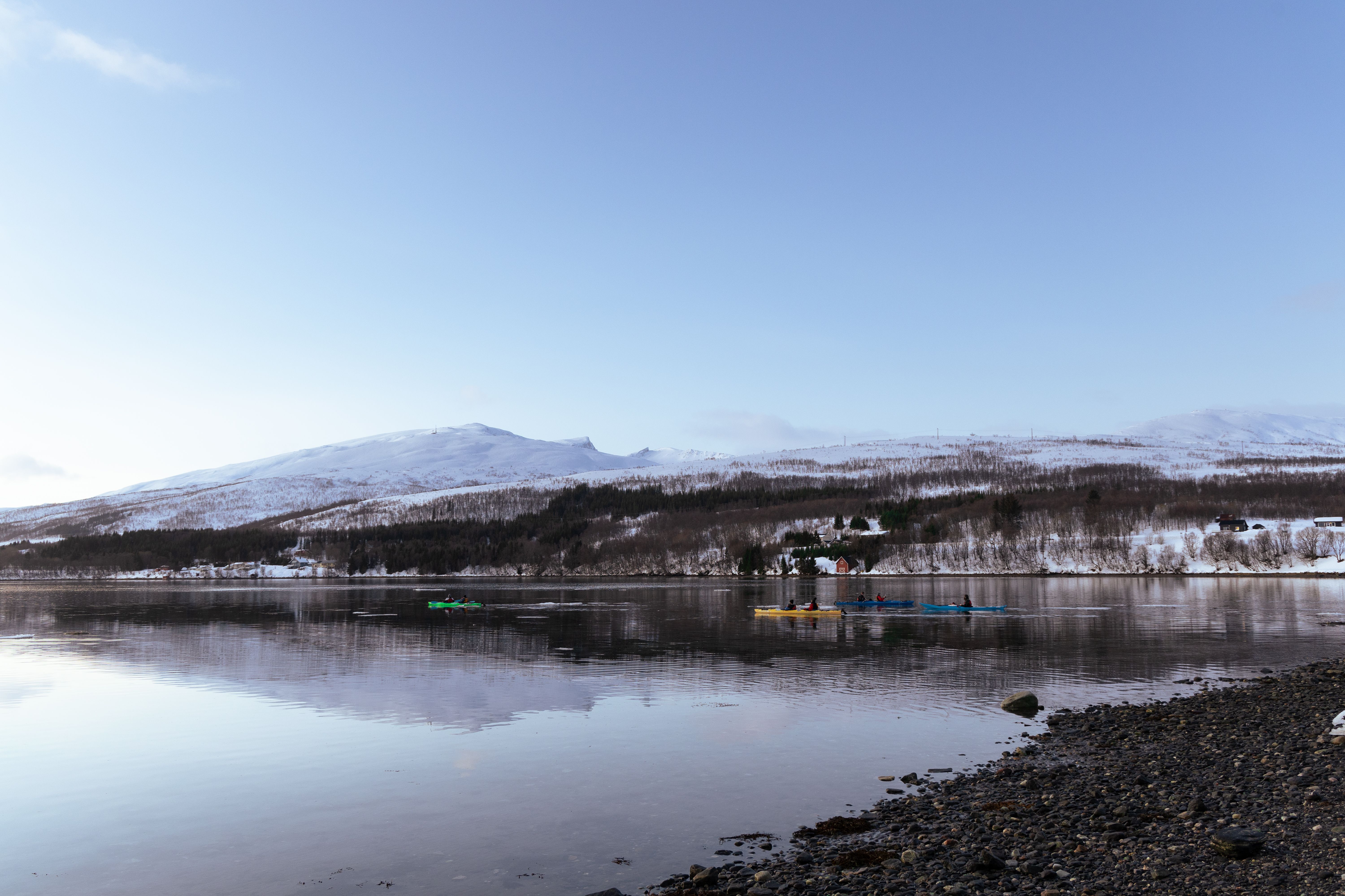 Kayakers paddle on a calm lake with snow-covered mountains in Norway during winter.