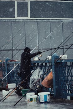 A person fishing on a snowy urban bridge surrounded by fishing gear.