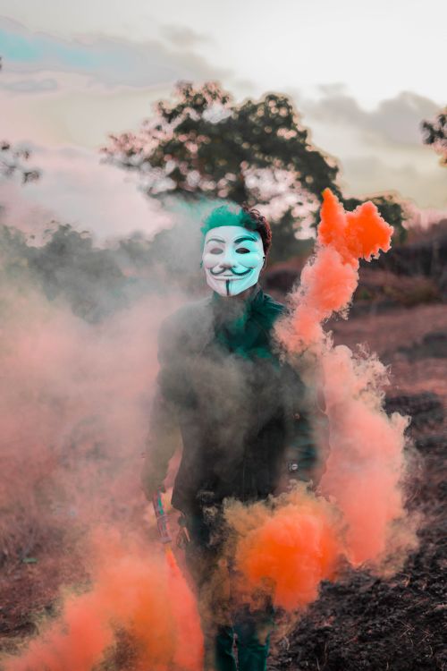 Free Person wearing a Guy Fawkes mask surrounded by colorful smoke in an outdoor setting. Stock Photo