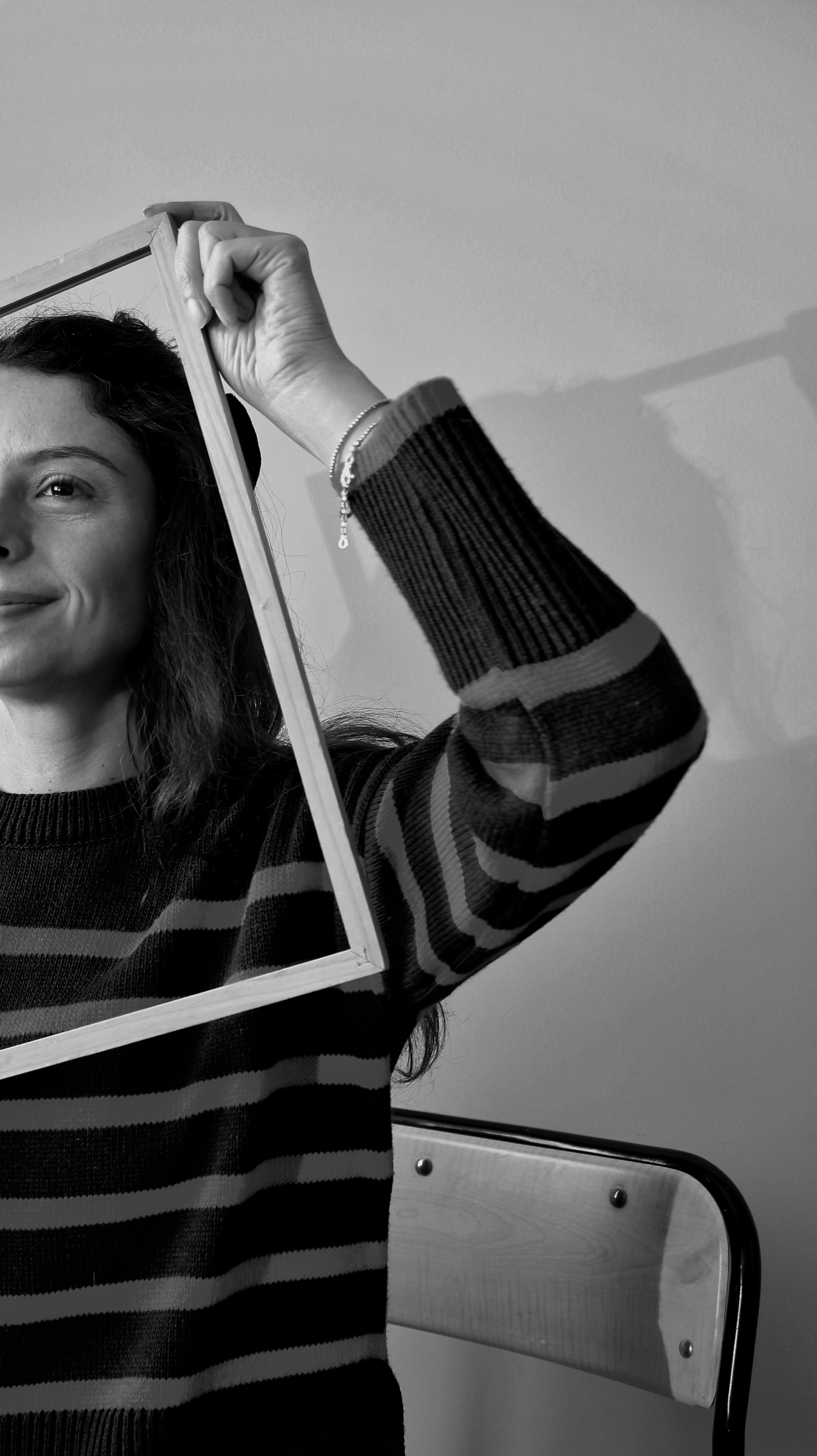 Black and white portrait of a woman playfully framing her face with a wooden frame.