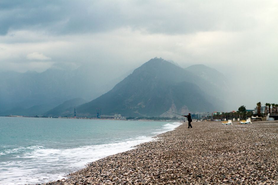 A lone fisherman on a pebble beach with majestic mountains under a cloudy sky, offering a serene vibe.