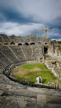 Ruins of a Roman amphitheater with a crane and cloudy sky, showcasing ancient architecture.