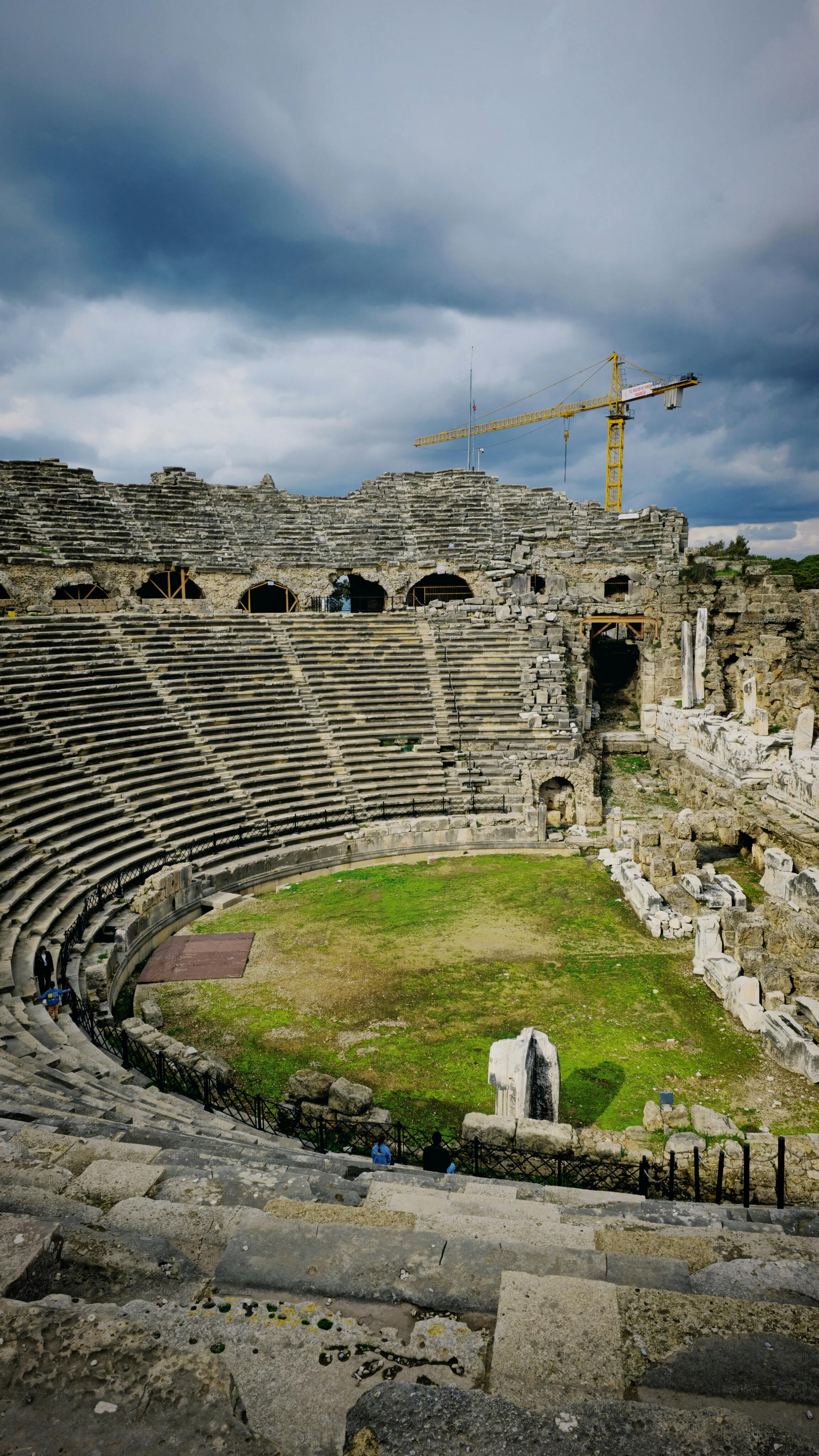 Kostenlos Ruinen eines römischen Amphitheaters mit Kran und bewölktem Himmel, die antike Architektur zeigen. Stock-Foto