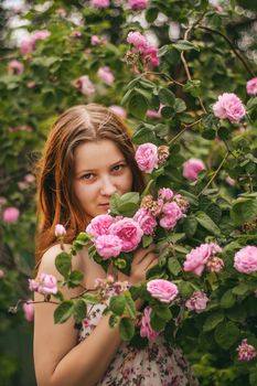 A young woman in a floral dress enjoying pink roses in a lush garden setting.