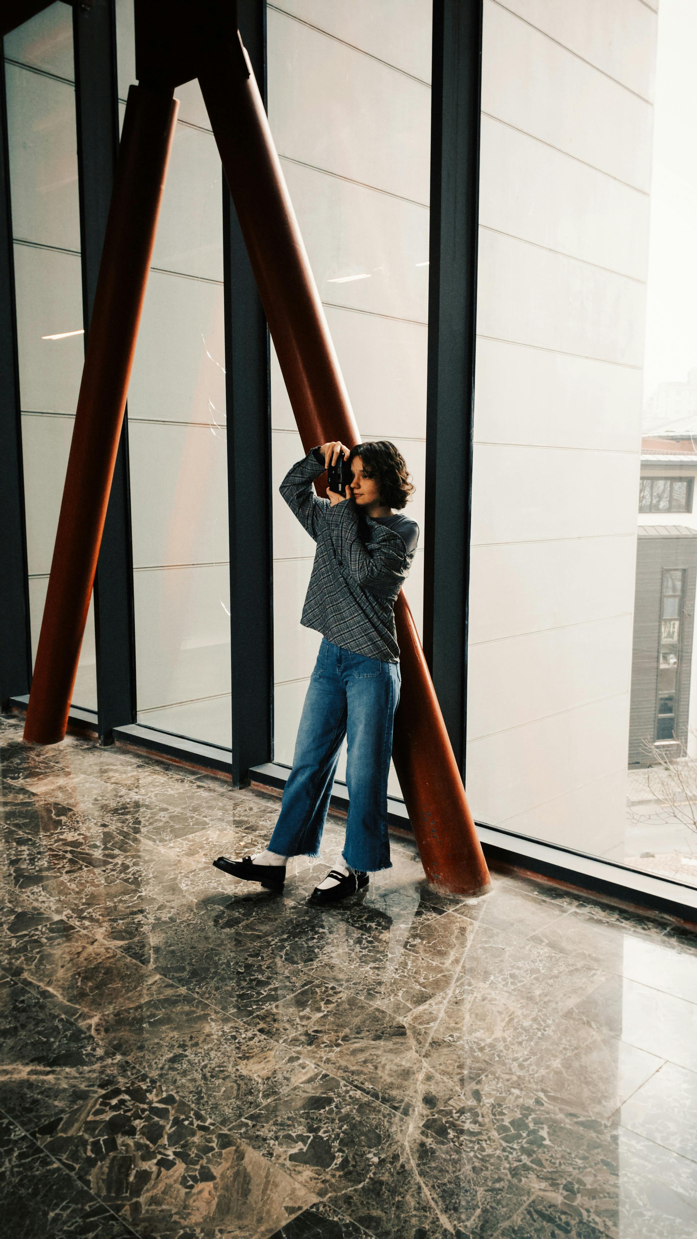 Stylish woman posing against a modern building window, reflecting urban life.