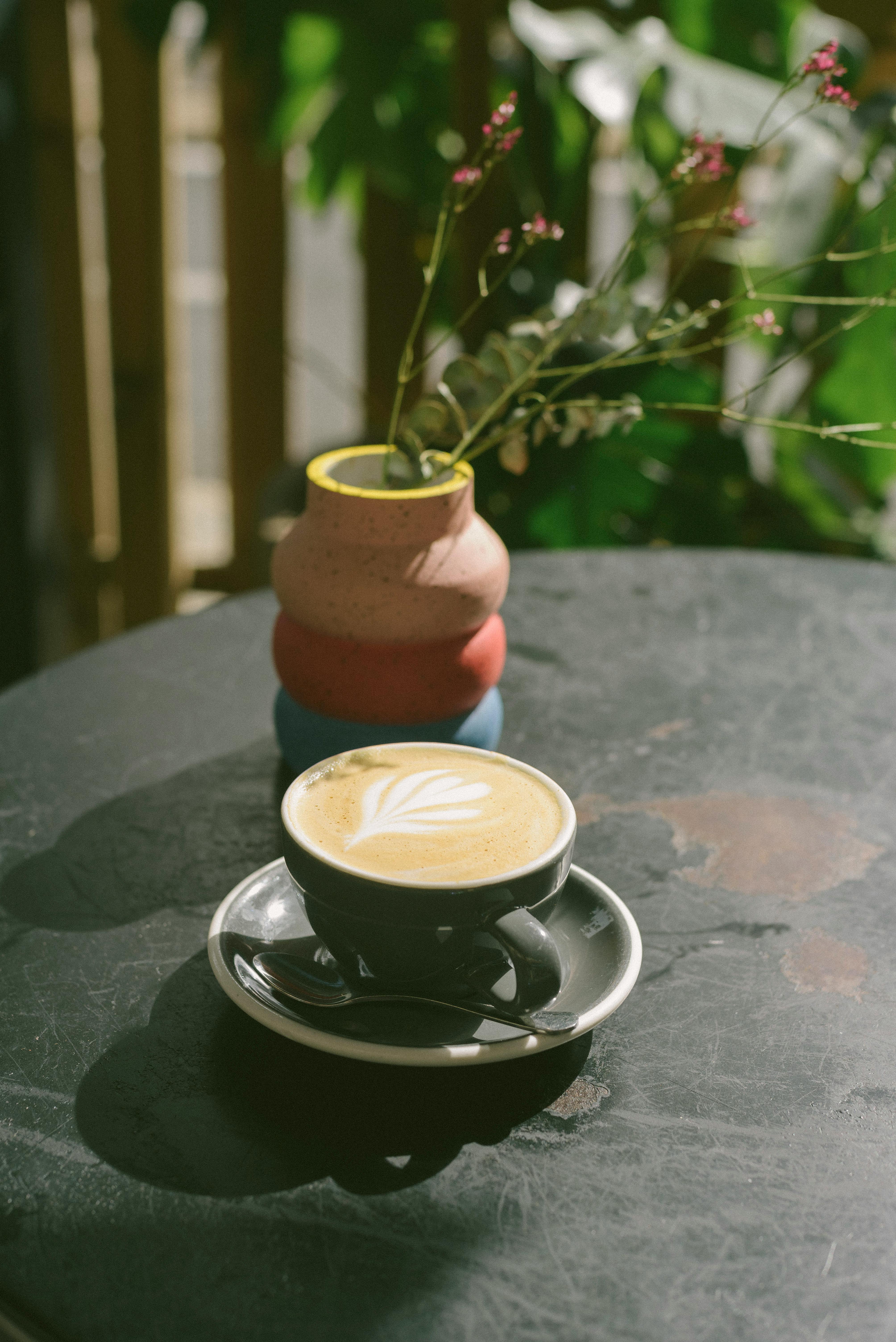 A latte in a black cup with latte art beside a colorful vase on an outdoor table.