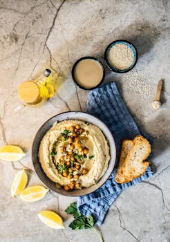 Flat lay of hummus with chickpeas, tahini, olive oil, lemon wedges, and bread slices.