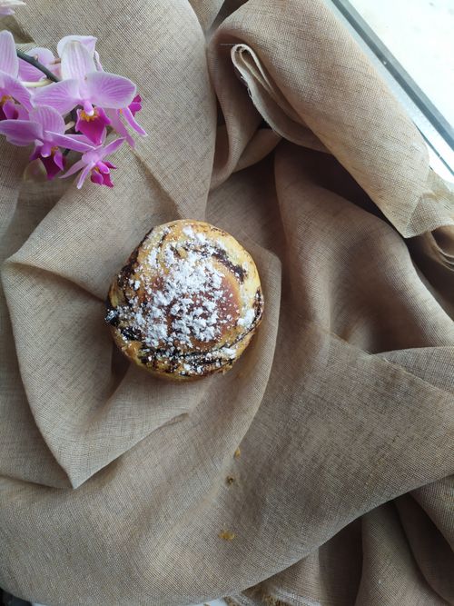 Free Cinnamon roll sprinkled with icing sugar next to a pink orchid on beige fabric. Stock Photo