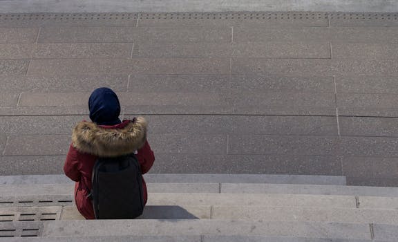 A person in a red coat sits alone on concrete steps, contemplating in an urban setting.