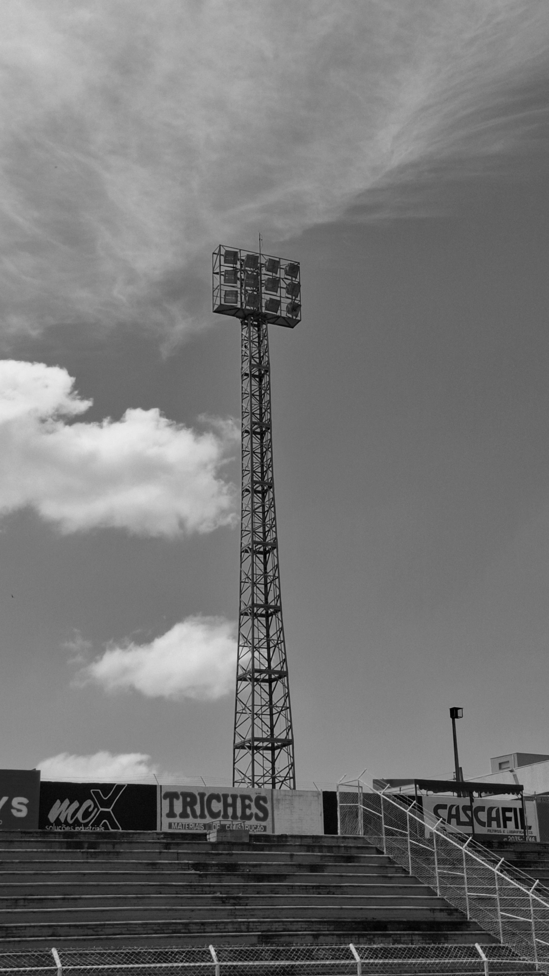 Black and white photo of a tall stadium floodlight tower under a cloudy sky.