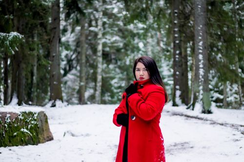 Free A woman in a vibrant red coat stands in a snow-covered forest during winter, creating a striking contrast. Stock Photo