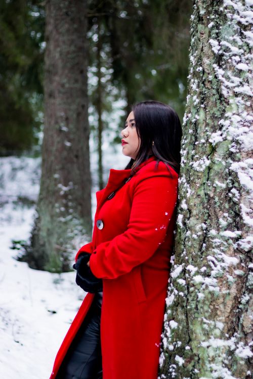 Free A woman in a red coat leans against a tree in a snowy forest, lost in thought. Stock Photo