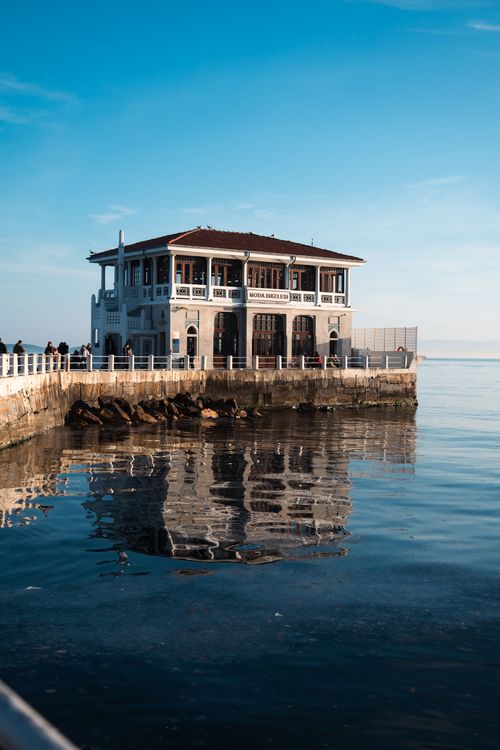 Free A scenic view of a waterfront house reflecting in calm ocean waters, under a clear blue sky. Stock Photo