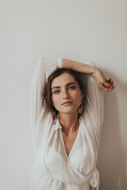 Foto De Mujer Con Blusa Blanca De Manga Larga Y Cuello En V Posando Junto A La Pared Blanca Con La Mano Sobre La Cabeza