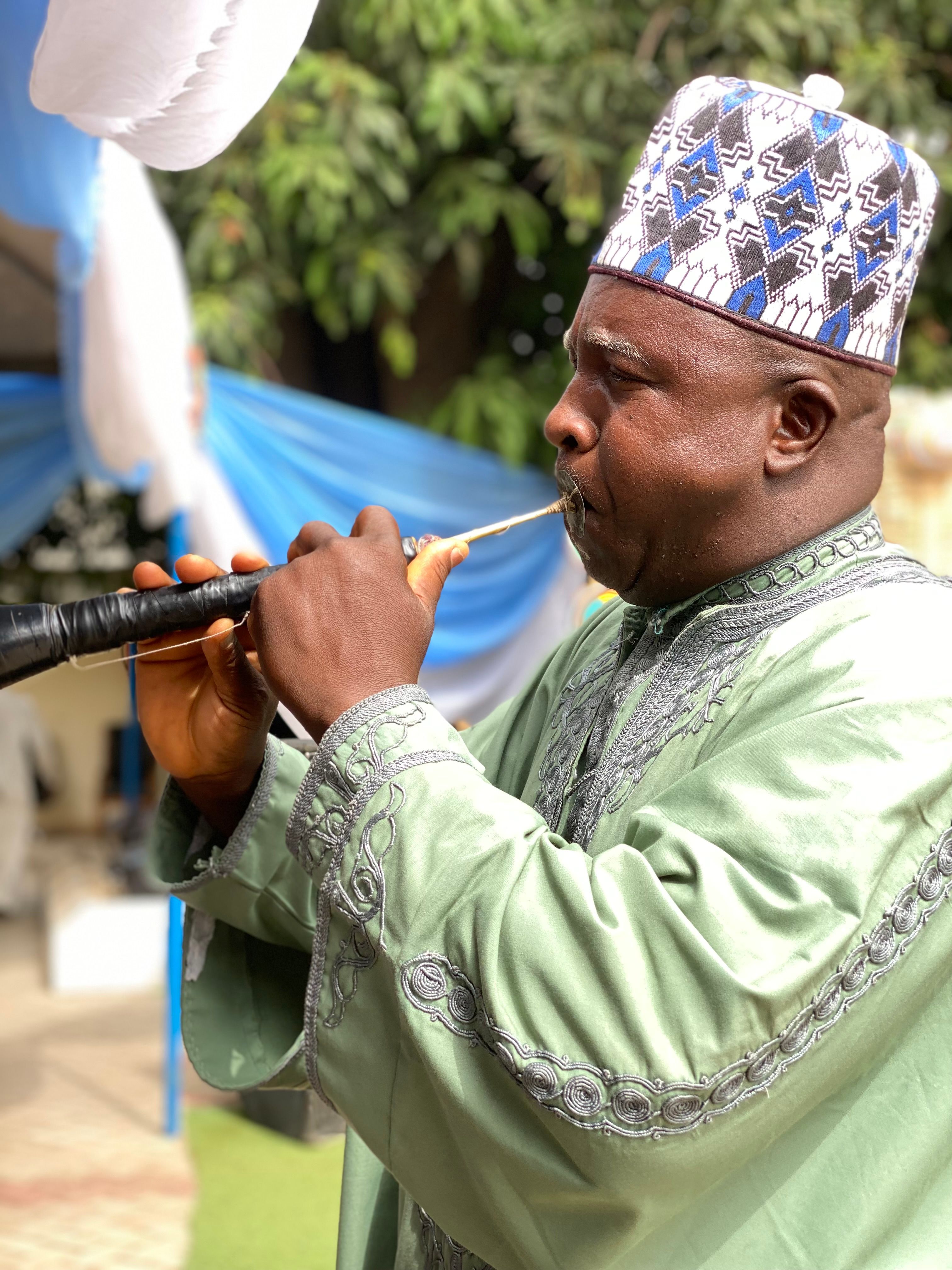 African man in traditional clothing playing a wind instrument at an outdoor event.