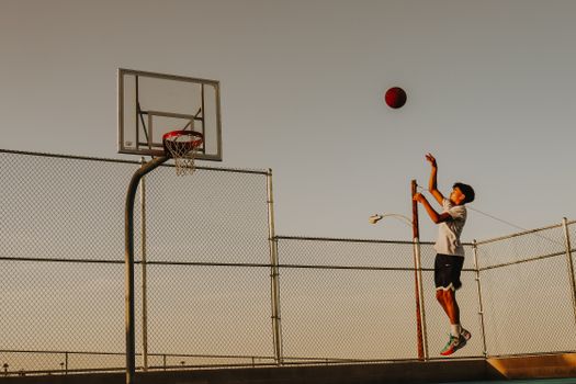 Teen boy playing basketball outdoors, shooting at hoop with sun setting.