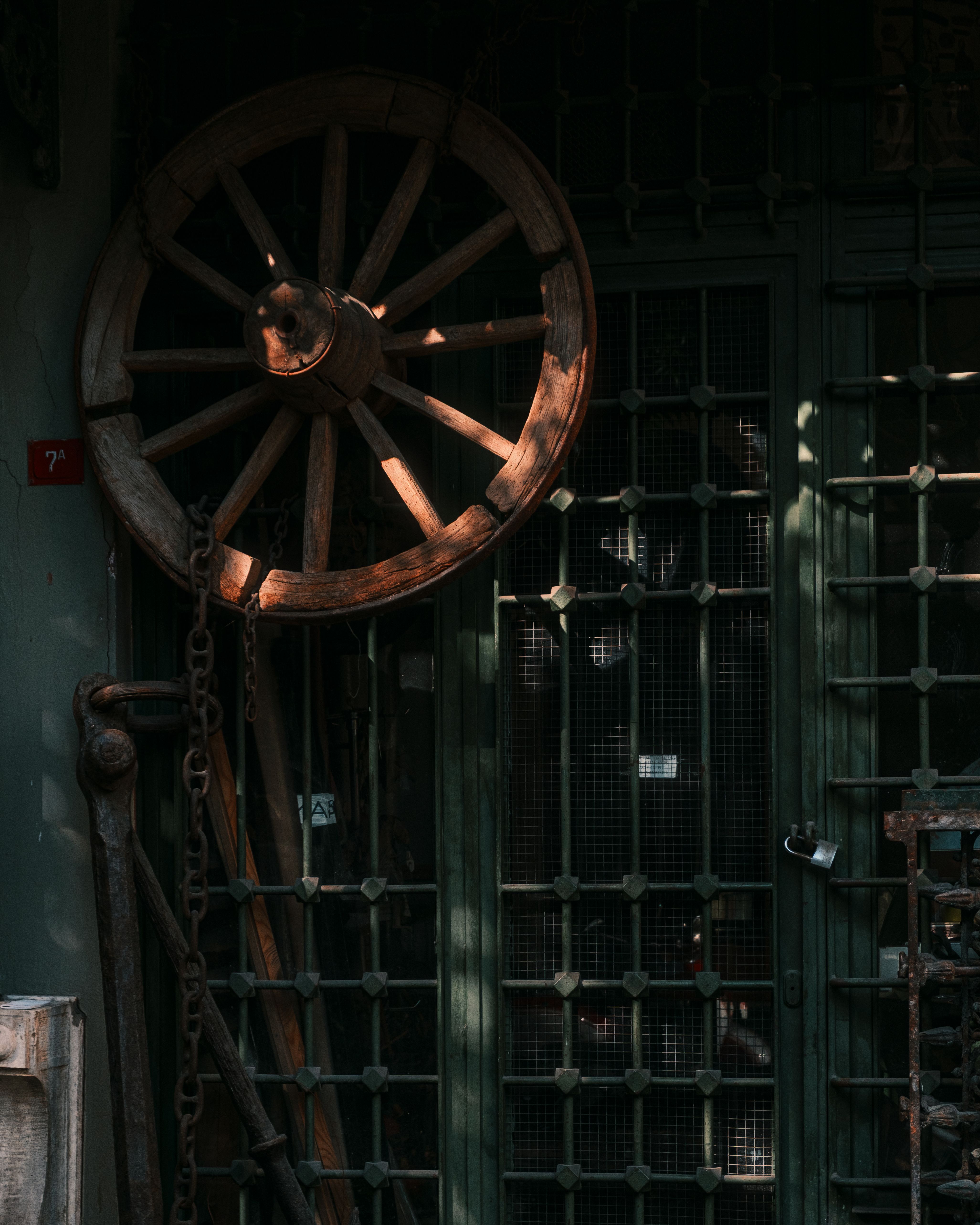 A rustic wooden cartwheel leaning against a metal grated door in dim lighting.
