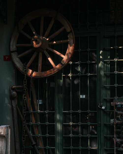 Free A rustic wooden cartwheel leaning against a metal grated door in dim lighting. Stock Photo