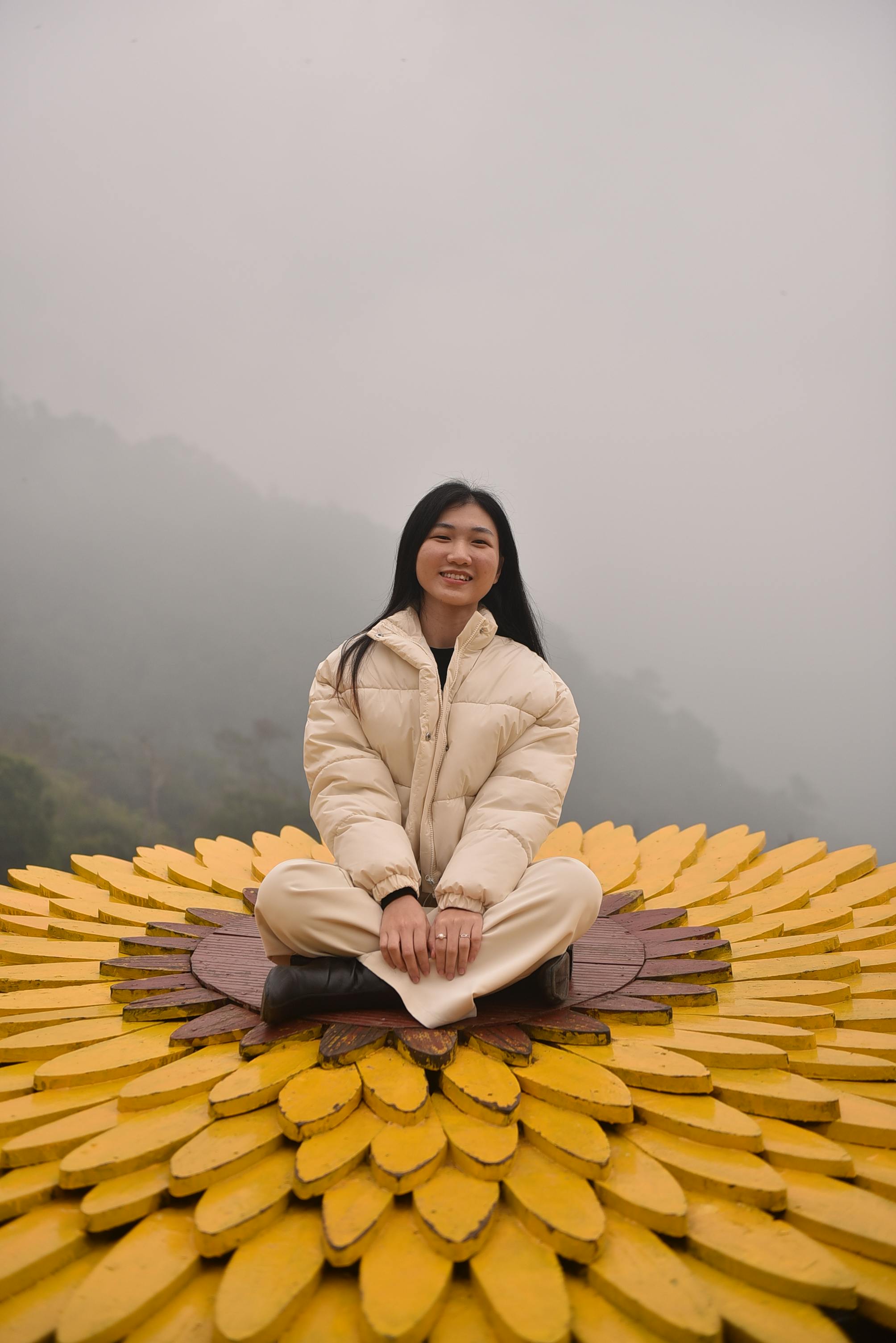 Young woman in a beige coat sits on a large yellow flower sculpture against a foggy landscape.