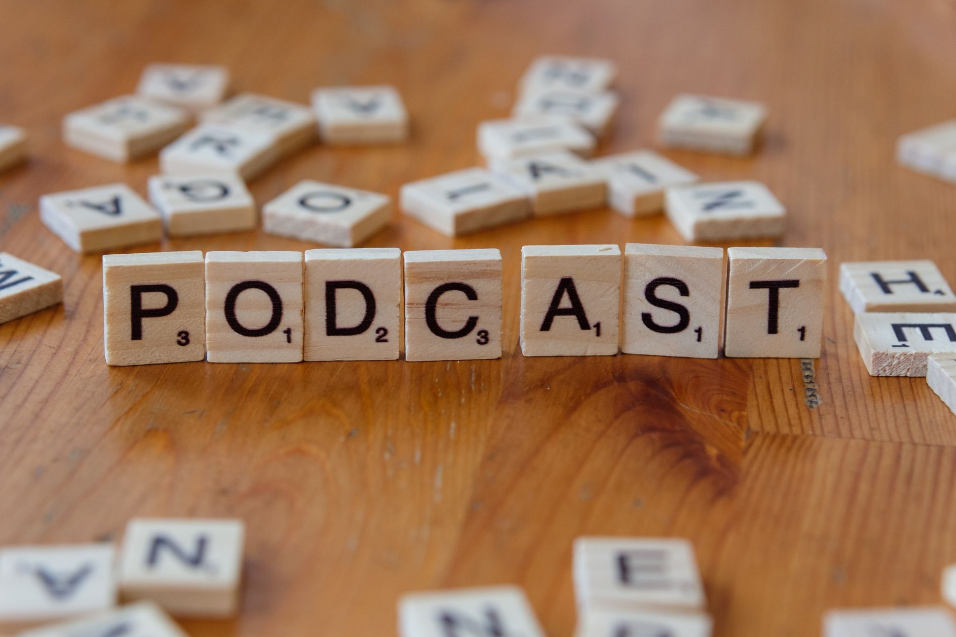 Scrabble tiles arranged to spell 'PODCAST' on a wooden surface with scattered letters.
