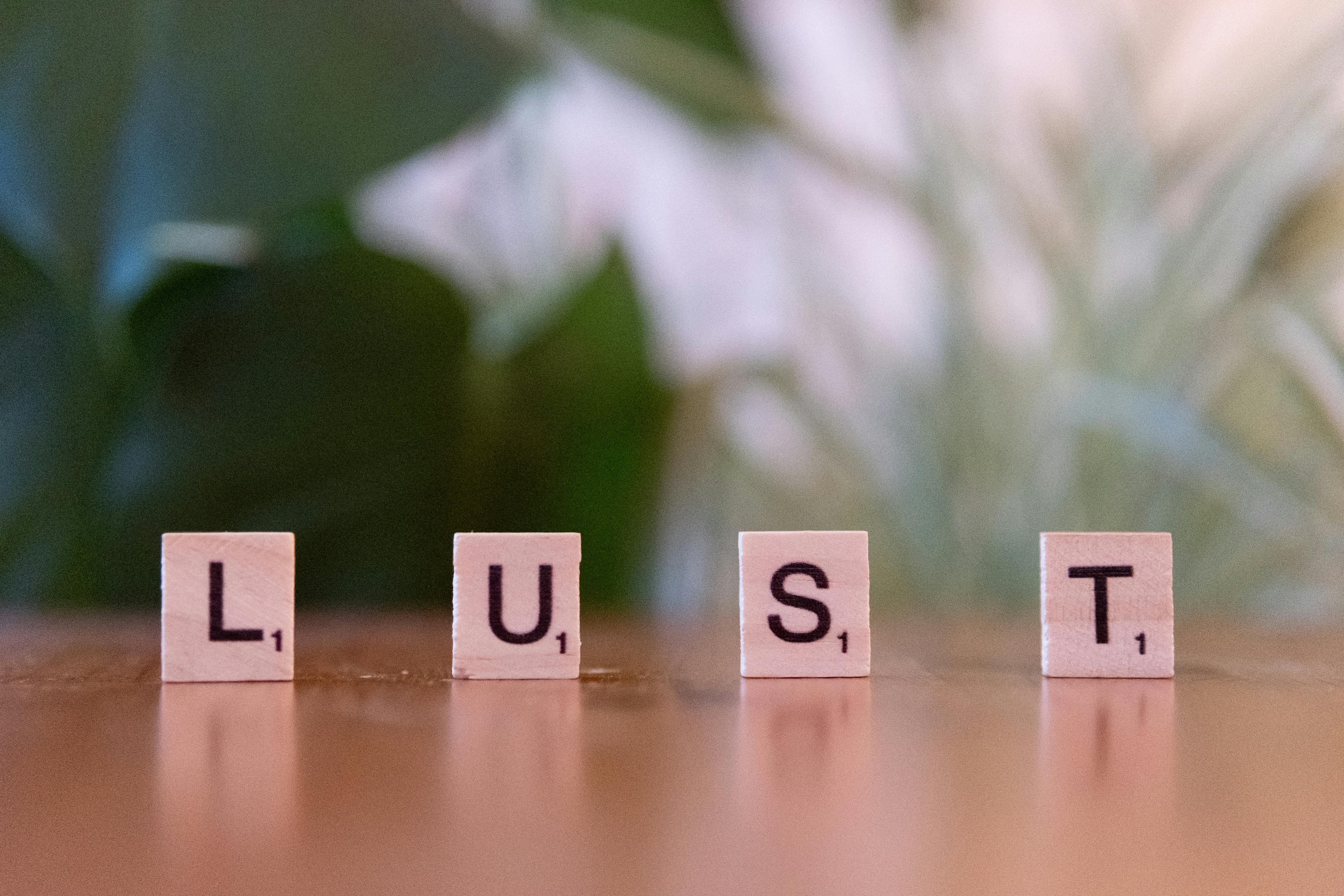 Scrabble tiles spelling 'LUST' on a wooden surface with a blurred green background.