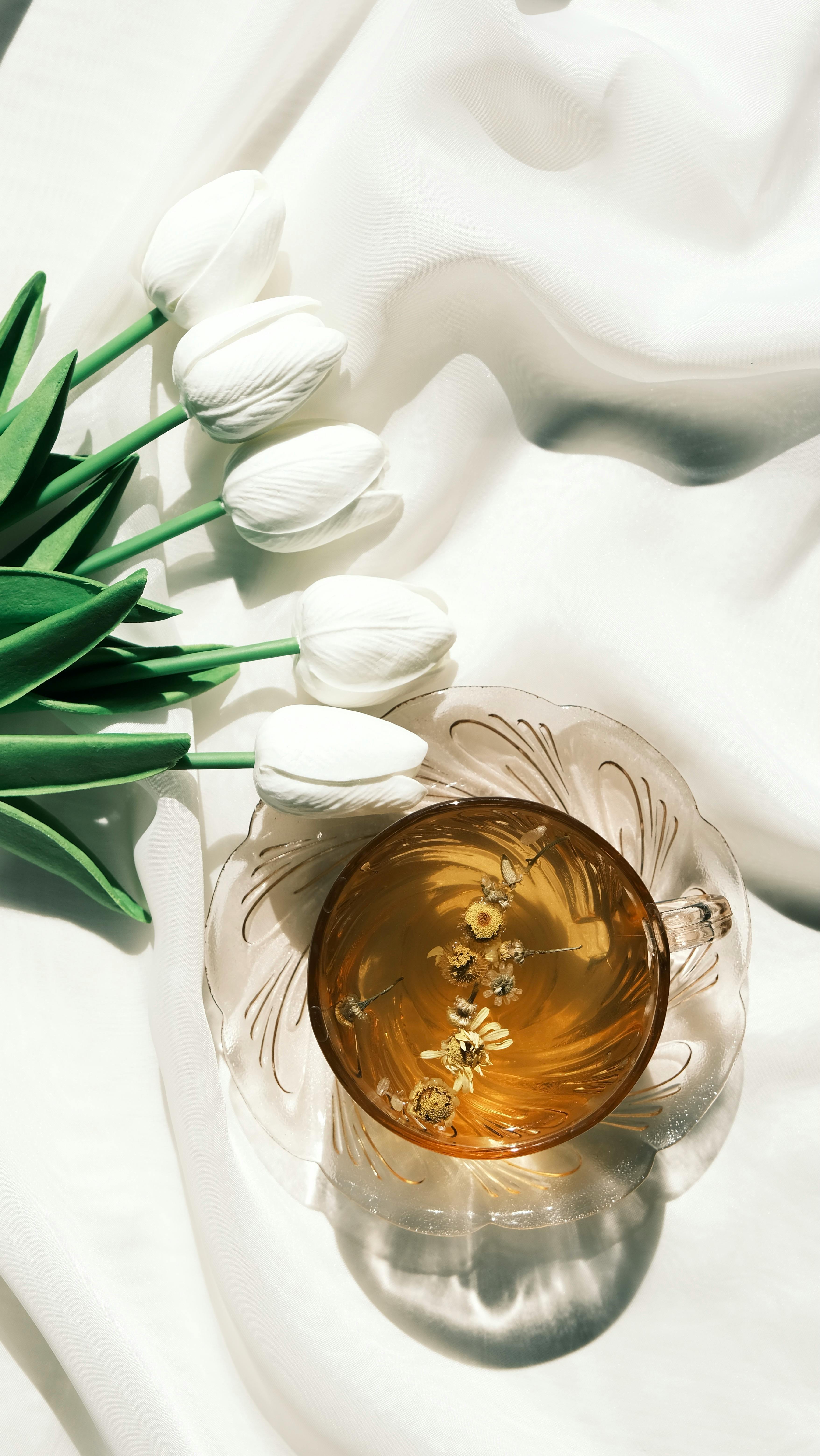 A delicate tea cup with chamomile on fabric beside white tulips in soft sunlight.