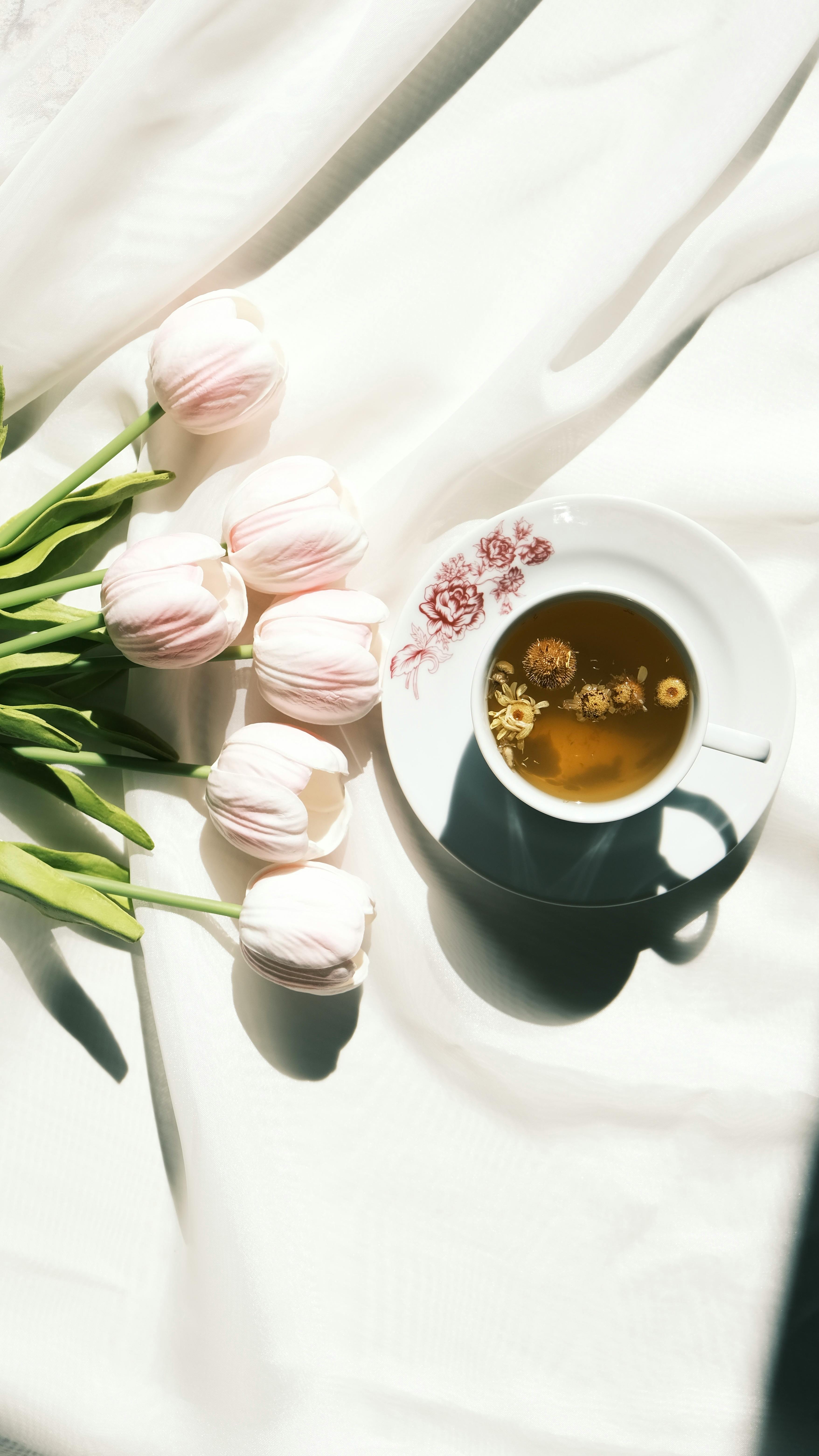 A floral tea cup and tulips beautifully lit by sunlight on a soft fabric surface.