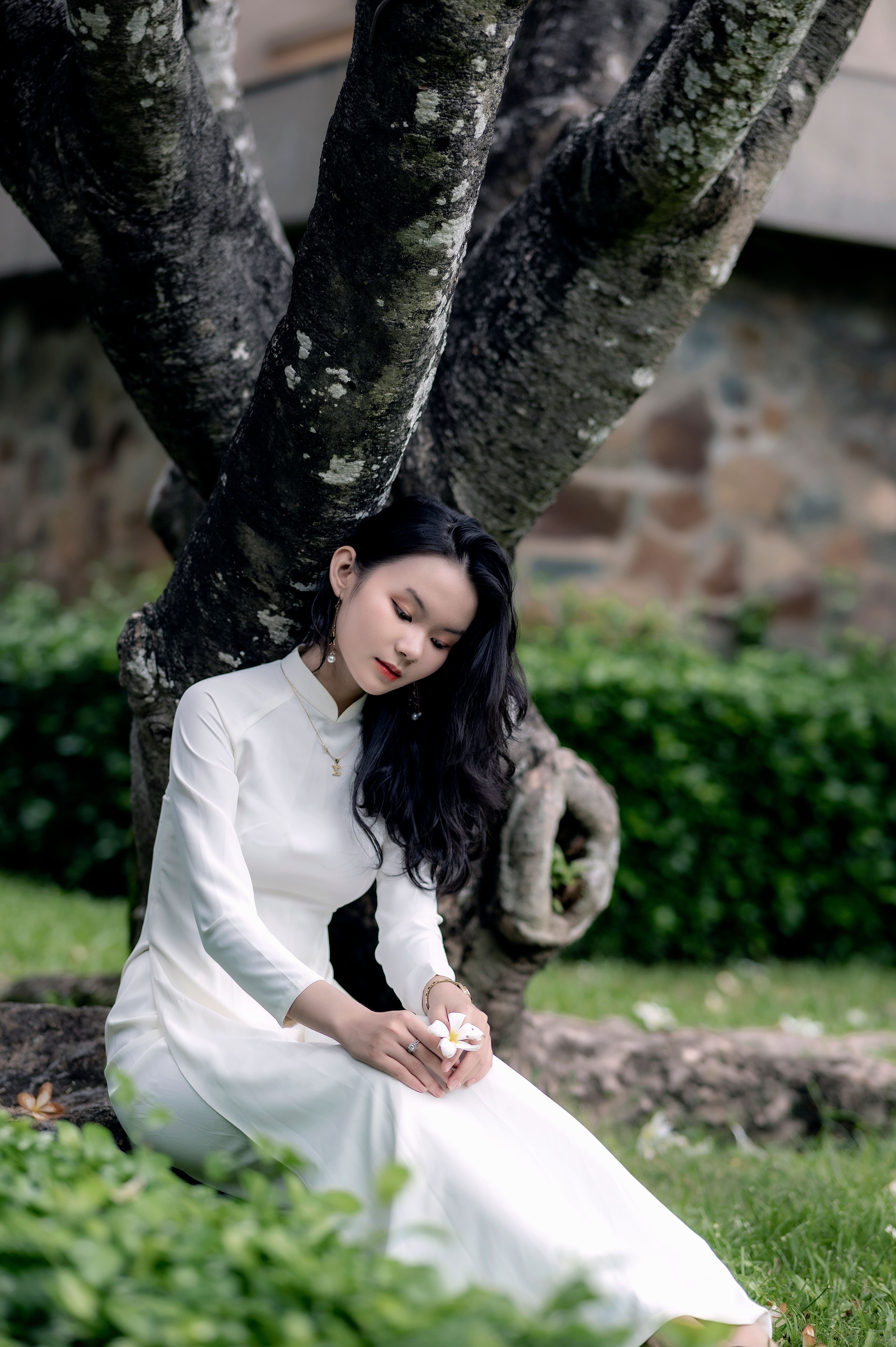 Elegant woman in white dress sits thoughtfully by a large tree outdoors.