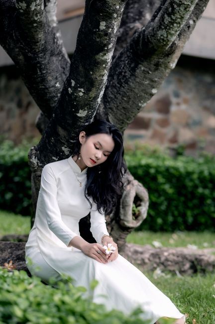 Elegant woman in white dress sits thoughtfully by a large tree outdoors.