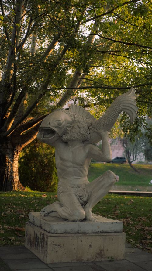 Free Stone sculpture depicting a mythical sea creature holding a fish, set in a serene park. Stock Photo