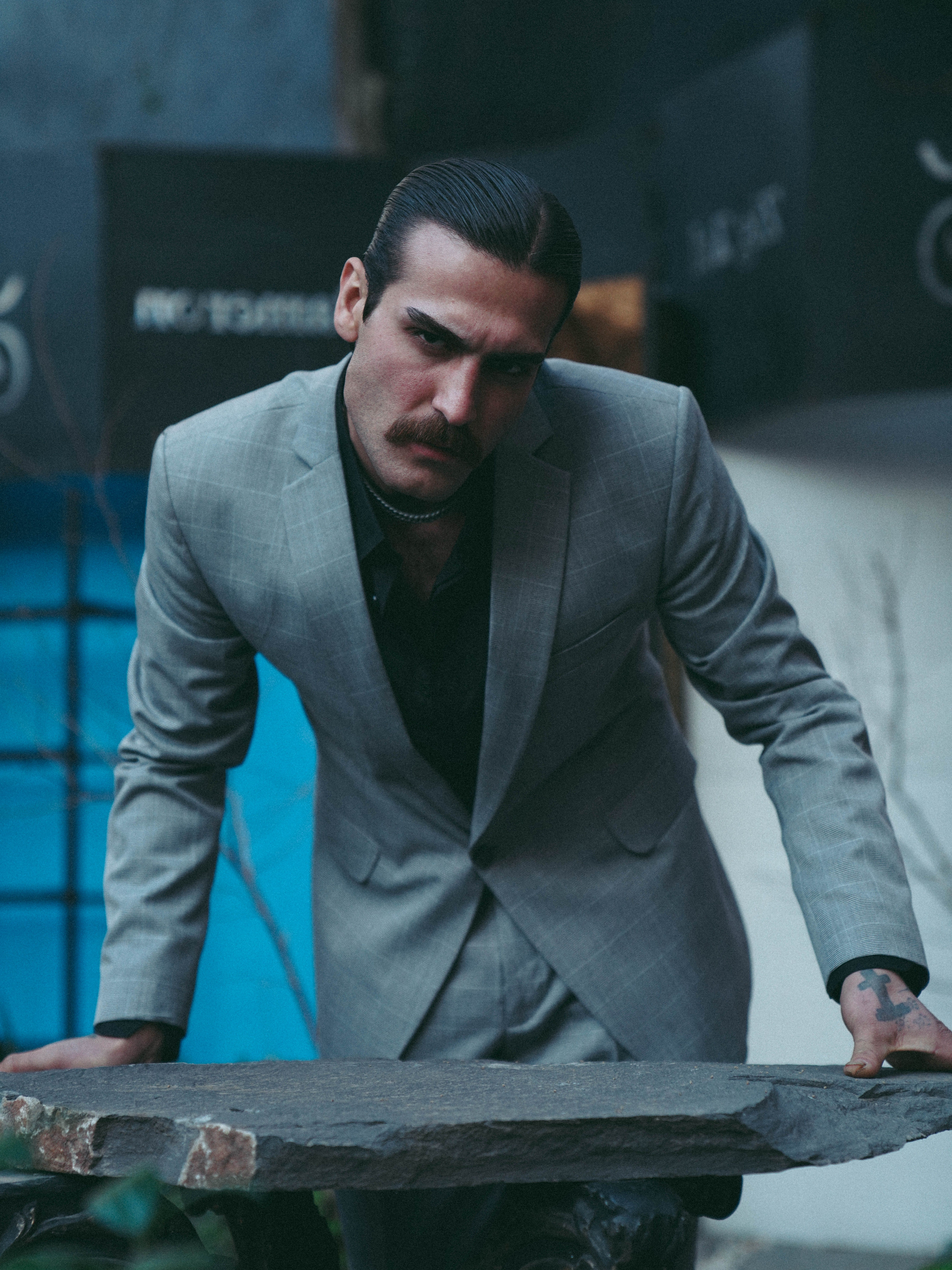 A man in a gray suit leans confidently forward, exuding elegance and charm in an urban setting.