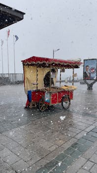 Red street food cart during snowfall in an urban setting, offering a cozy winter vibe.